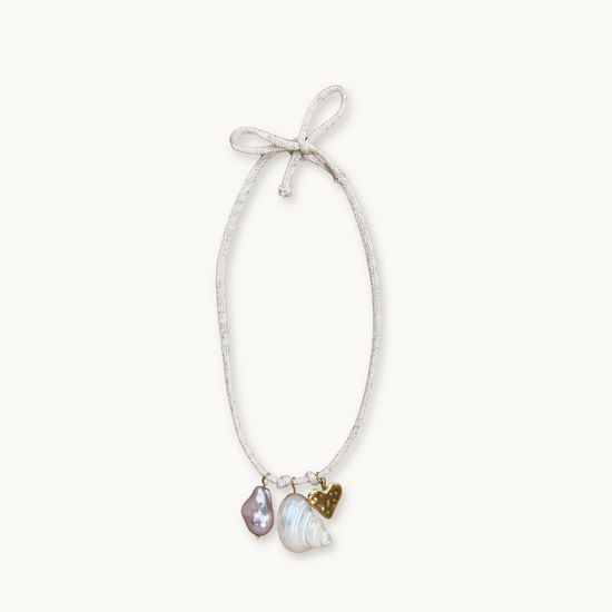 A necklace with a thin, textured white cord tied in a bow features three pendants: a purple pearl, a gold heart, and a seashell. Elegant, beach-inspired.