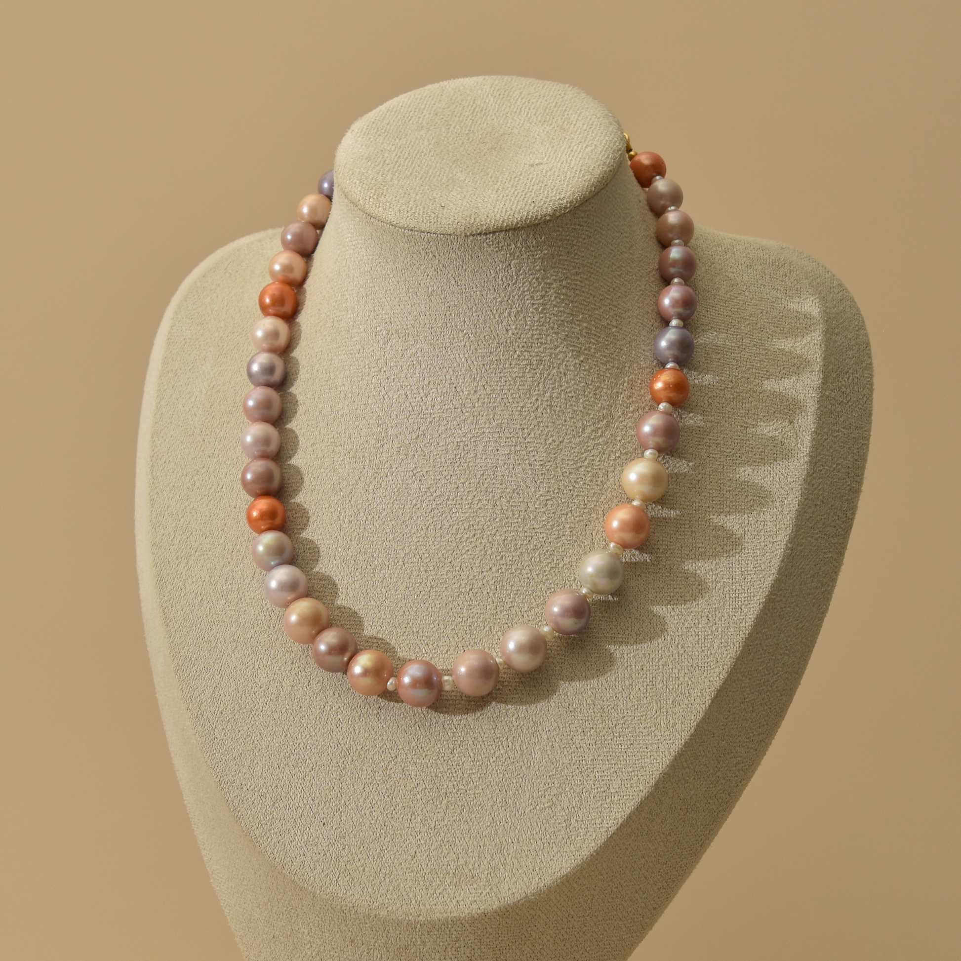 A strand of irregular freshwater pearl necklace in soft pastel colors, displayed on a beige fabric jewelry bust against a warm beige background.
