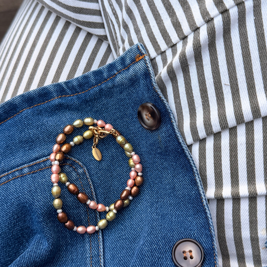 A close-up of a pearl necklace with pink, green, and brown pearls and gold-toned metal, resting on a blue denim jacket over a black and white striped shirt.