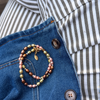 A close-up of a pearl necklace with pink, green, and brown pearls and gold-toned metal, resting on a blue denim jacket over a black and white striped shirt.