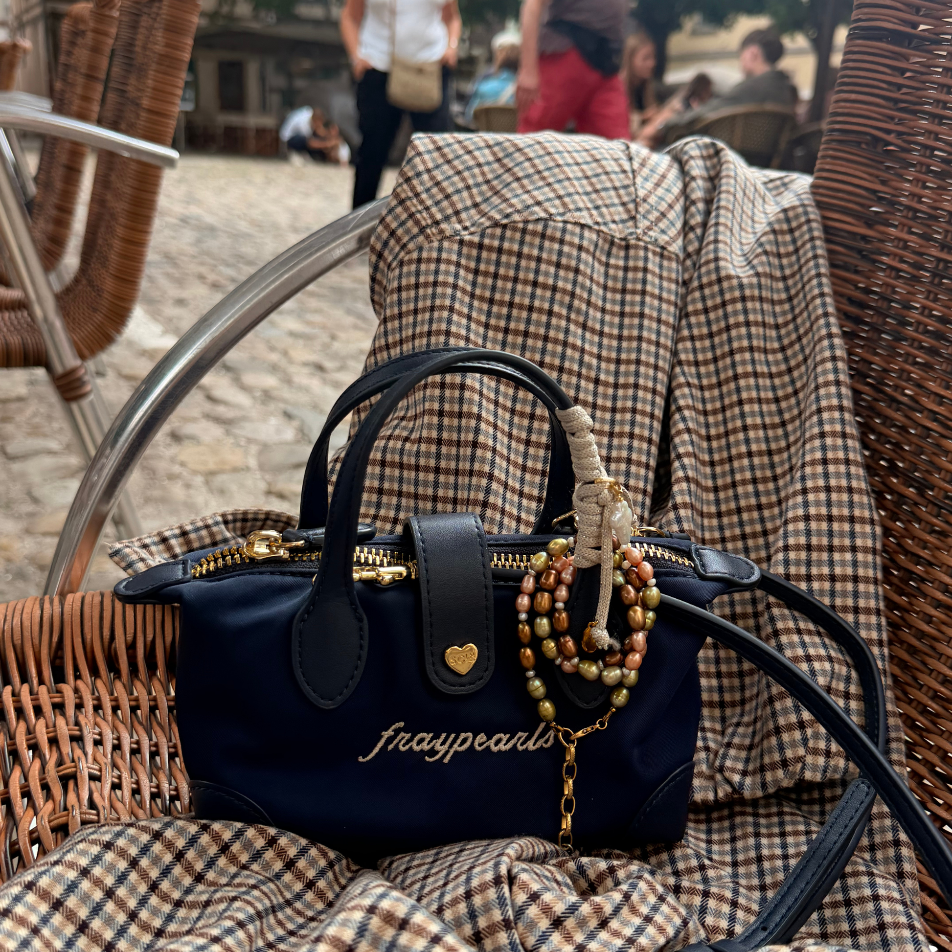 A navy blue handbag with gold hardware and the brand name 'fraypearls' embroidered on the front, resting on a brown and beige plaid blanket draped over a wicker chair in an outdoor cafe setting.