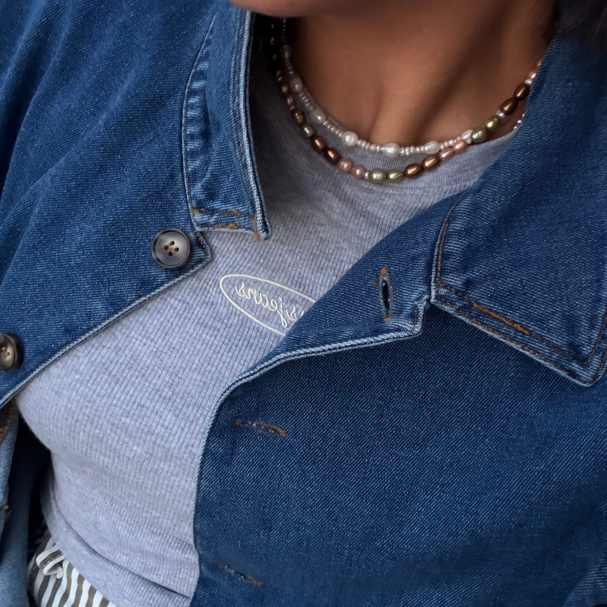Close-up of a person wearing a light gray ribbed t-shirt with a white embroidered logo, layered under a dark blue denim jacket, accessorized with a pearl necklace.