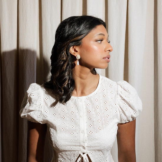 Woman wearing a white lace top against a neutral curtain background wearing ornate style bridal statement earrings. 