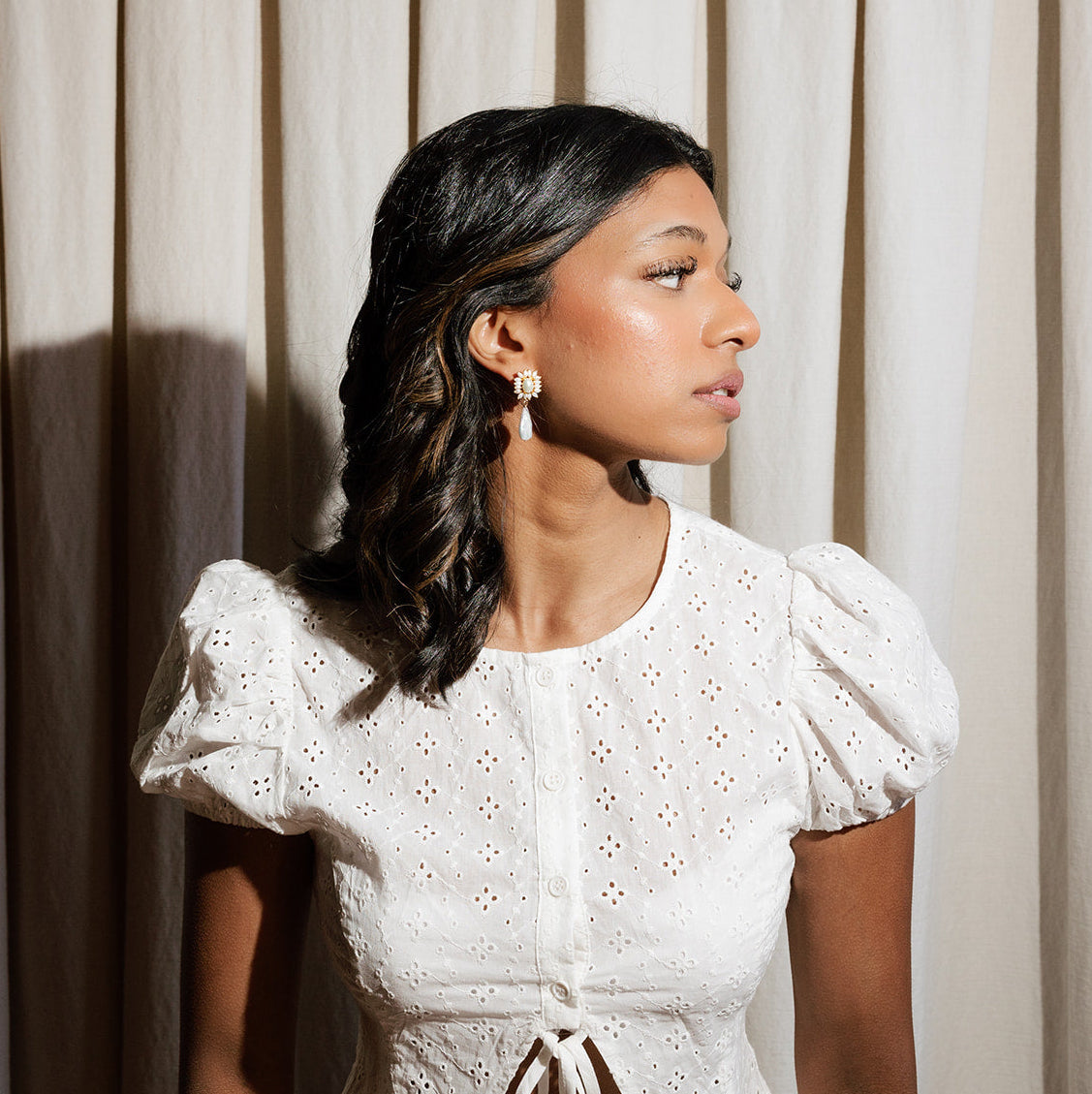 Woman wearing a white lace top against a neutral curtain background wearing ornate style bridal statement earrings. 