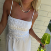 Woman in a white sundress with a pearl necklace, holding a green wine glass, stands against a light brick wall. The mood is casual and summery.