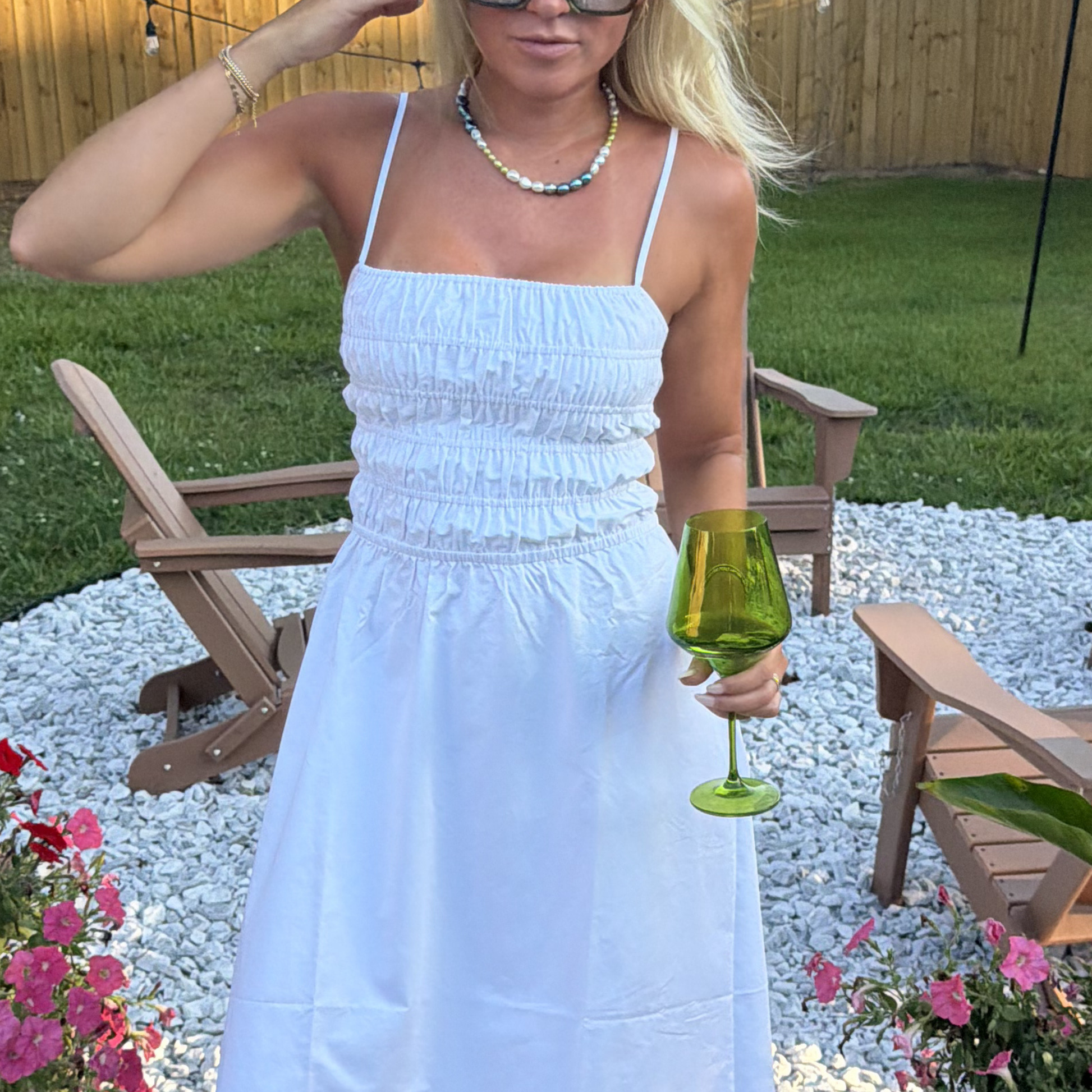 A person in a white dress holds a green wine glass in a backyard. They wear sunglasses and colorful pearl necklace, with wooden chairs and green grass behind.