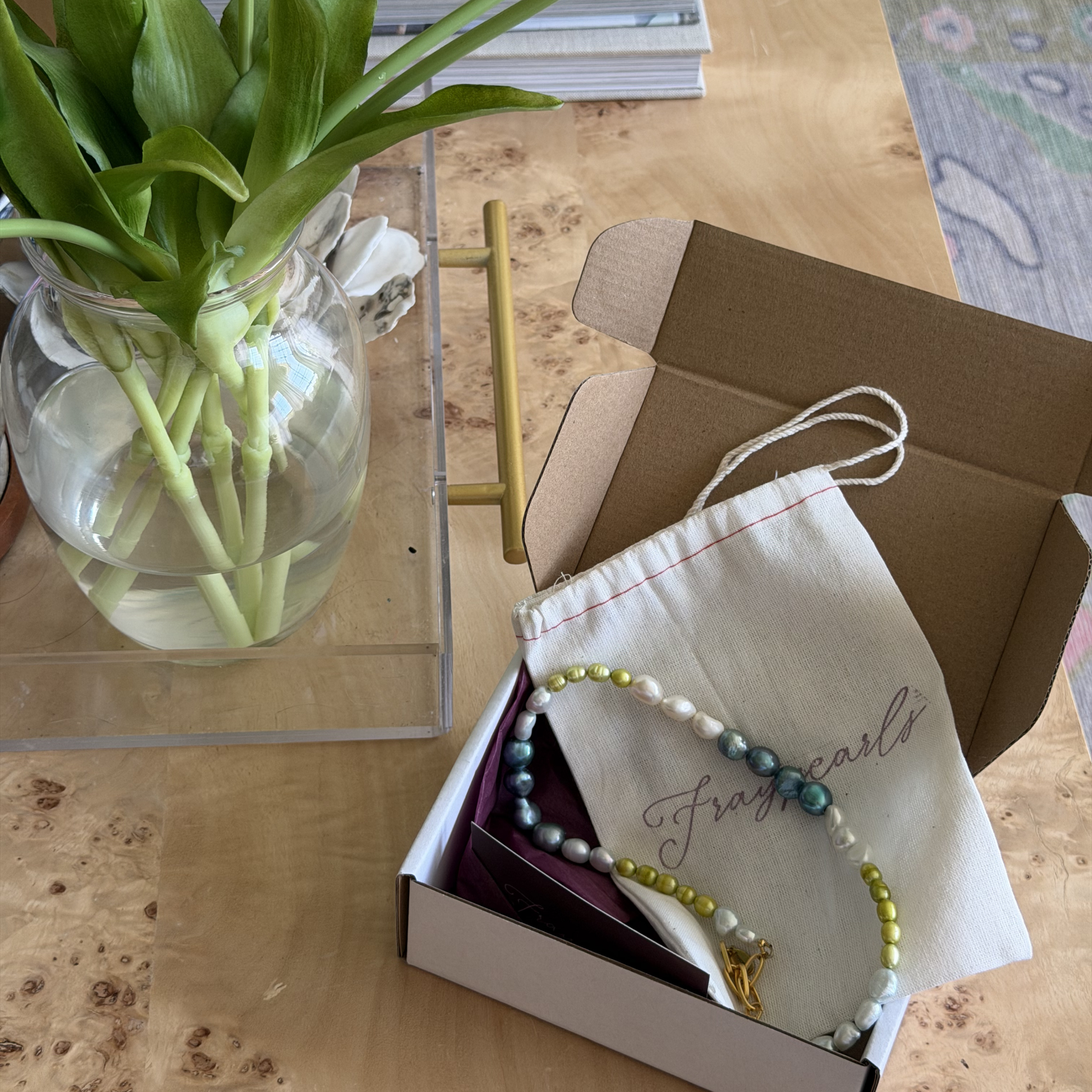 A pearl necklace with vibrant colors rests on a drawstring pouch inside an open cardboard box. Nearby, a vase holds fresh green tulips.
