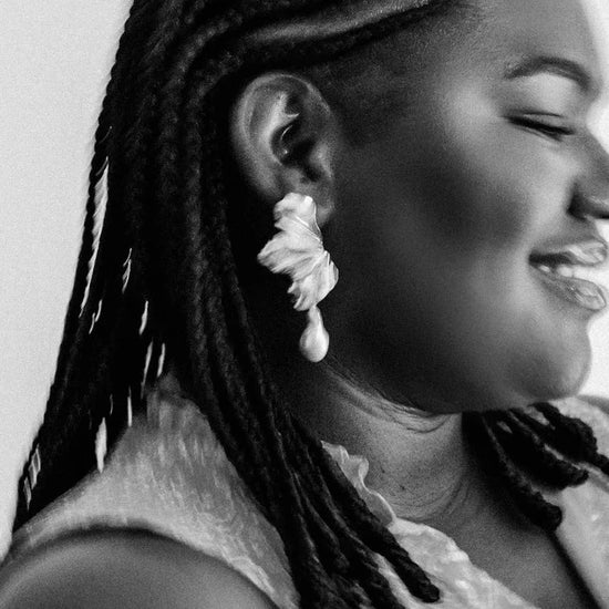 Black and white close-up of a person smiling, wearing braided hair and a large, intricate earring shaped like a flower. The mood is joyful and serene.