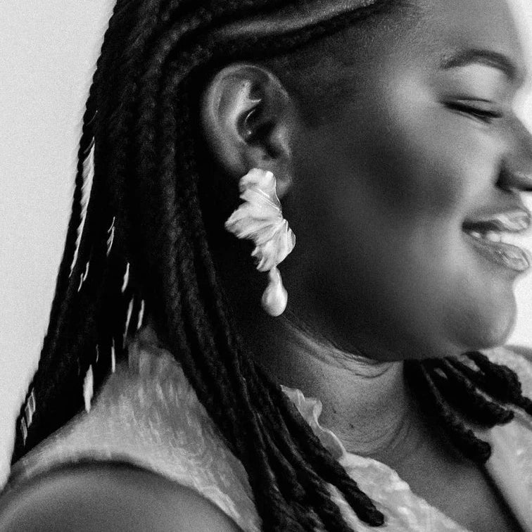 Black and white close-up of a person smiling, wearing braided hair and a large, intricate earring shaped like a flower. The mood is joyful and serene.