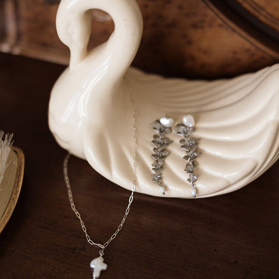 Elegant swan dish holds silver leaf earrings and pearl pendant necklace on dark wood surface, creating a vintage and sophisticated vibe.