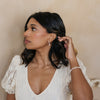 Woman in a white lace dress adjusting her hair against a neutral background in bridal wedding dress jewelry