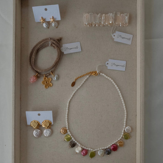 Assorted jewelry on a beige background featuring a pearl necklace with colorful pendants, gold shell earrings, beaded bracelets, and a clear hair clip.