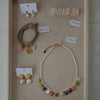 Assorted jewelry on a beige background featuring a pearl necklace with colorful pendants, gold shell earrings, beaded bracelets, and a clear hair clip.