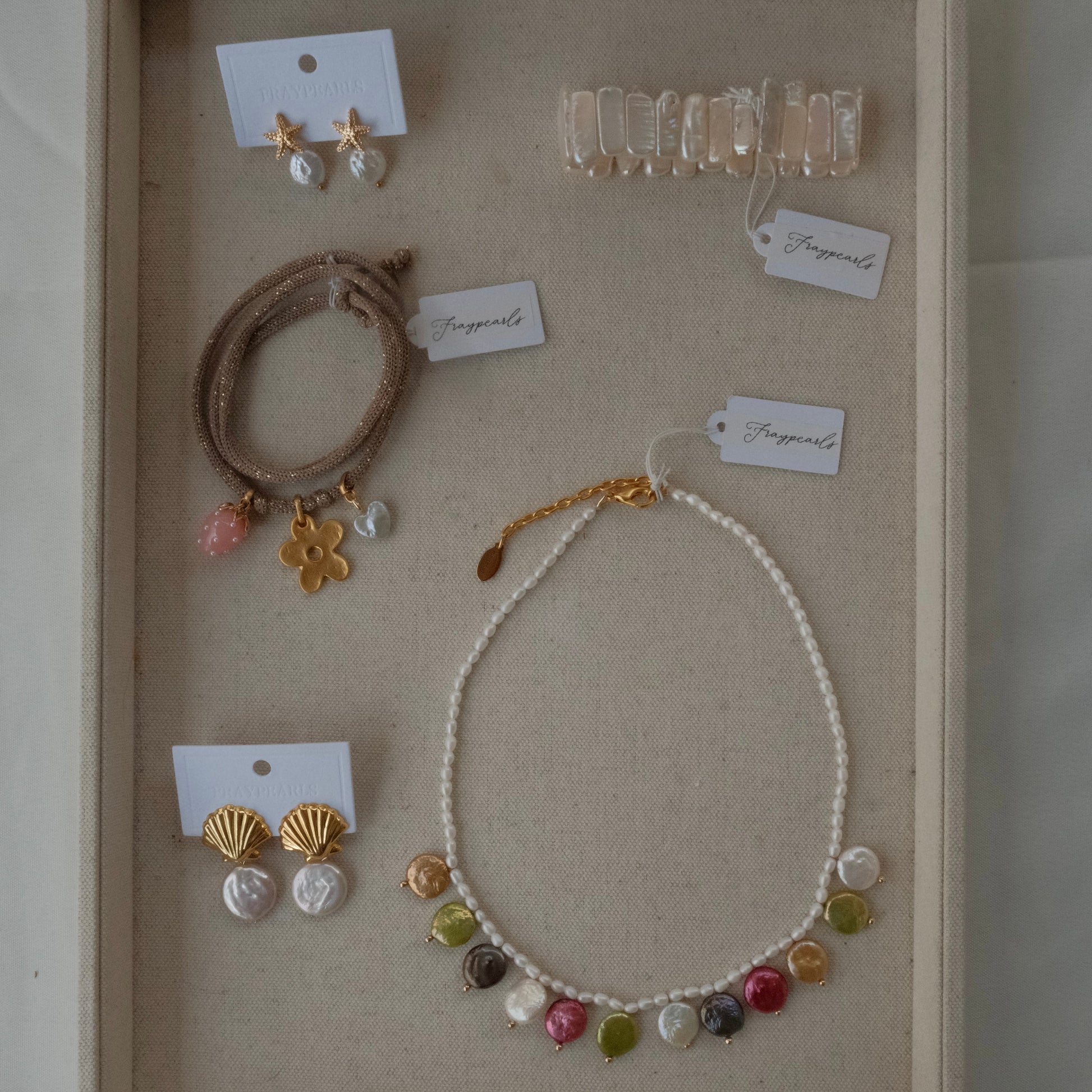 Assorted jewelry on a beige background featuring a pearl necklace with colorful pendants, gold shell earrings, beaded bracelets, and a clear hair clip.