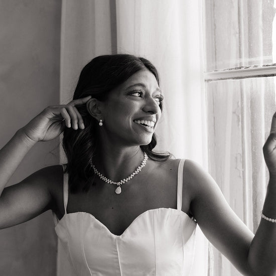 Woman in a white dress with jewelry, standing by a window. She’s wearing classic tailored bridal earrings and pearl necklace 