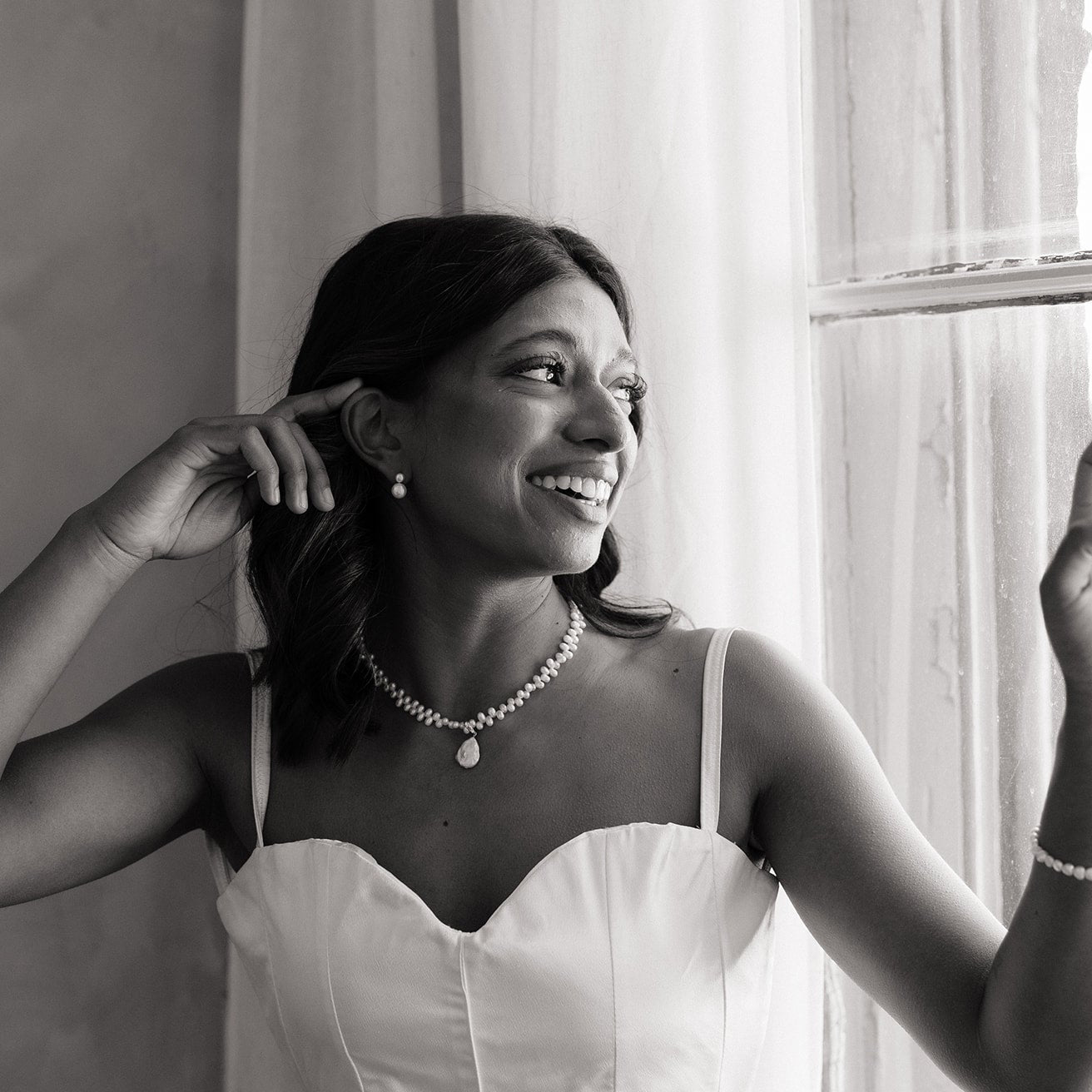 Woman in a white dress with jewelry, standing by a window. She’s wearing classic tailored bridal earrings and pearl necklace 