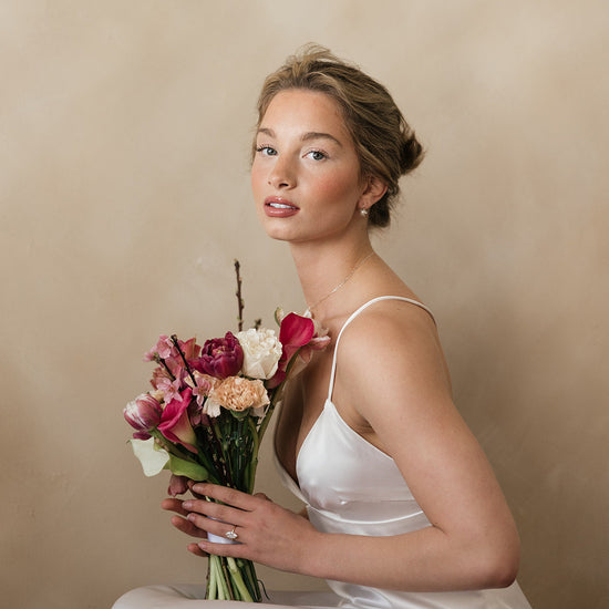 Woman in a white dress holding a bouquet of flowers against a beige background wearing wedding dress and bridal jewelry 