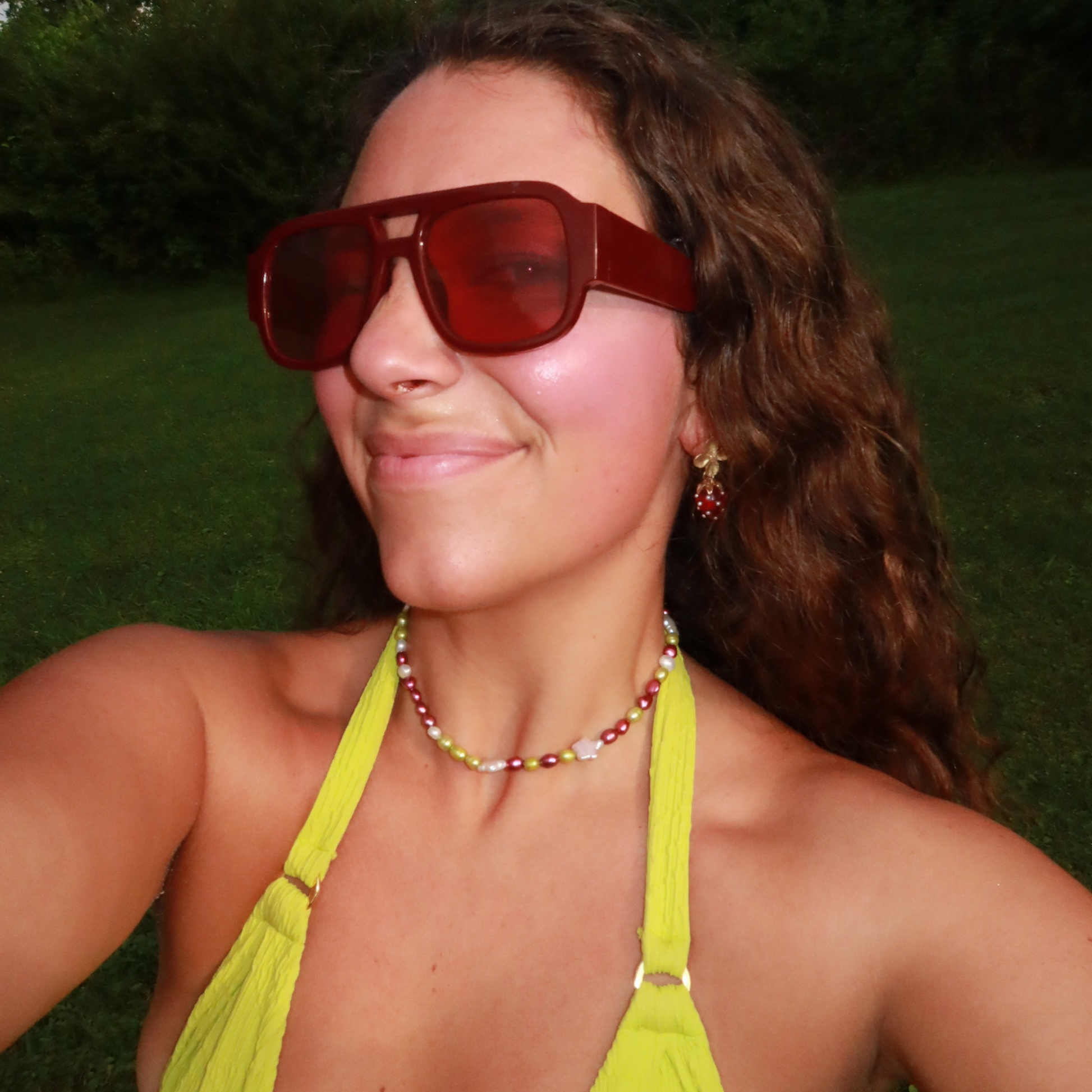 Woman smiling outdoors, wearing oversized red sunglasses, yellow top, colorful pearl necklace, and red earrings. The background is a grassy area.