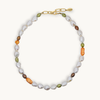 A necklace featuring baroque white, small olive green, brown, and orange pearls, accented with a gold chain clasp, shown on a light cream background.