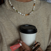 A close-up lifestyle shot of a person wearing a cream-colored knit turtleneck sweater and a pearl necklace, holding a white paper coffee cup with a black lid.