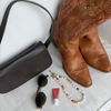 A styled flat-lay featuring a pair of tan leather cowboy boots with embroidered shafts, alongside a dark brown leather shoulder bag, black sunglasses, a red tube of lip balm, and a pearl beaded necklace on a white fabric background.