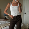 A woman takes a mirror selfie in a bedroom, wearing a white ribbed tank top and dark brown pants. She is accessorized with a pearl necklace, gold bracelets, and a watch.