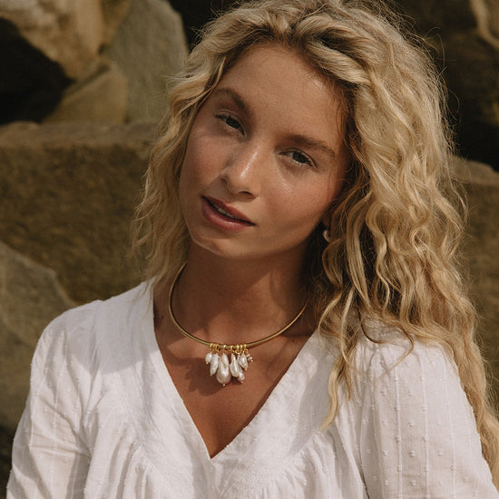 A woman with long blonde hair sits on a sandy beach in front of large rocks, wearing a white long-sleeve blouse with a ruffled V-neck and black shorts.
