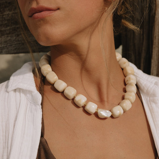 A close-up of a woman wearing a chunky cream-colored beaded necklace with a central iridescent pearl-like bead, styled with a white shirt and brown top.