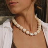 A close-up of a woman wearing a chunky cream-colored beaded necklace with a central iridescent pearl-like bead, styled with a white shirt and brown top.