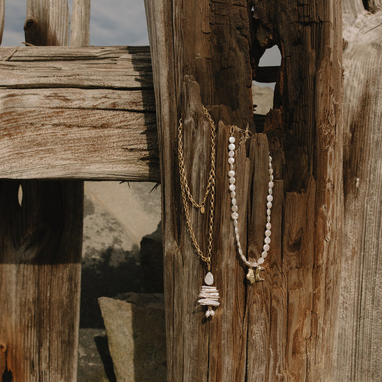 Two bohemian-style necklaces hanging from a rustic wooden fence post outdoors. One necklace has a gold chain with white pearls and a stacked white pearl pendant.