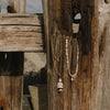 Two bohemian-style necklaces hanging from a rustic wooden fence post outdoors. One necklace has a gold chain with white pearls and a stacked white pearl pendant.