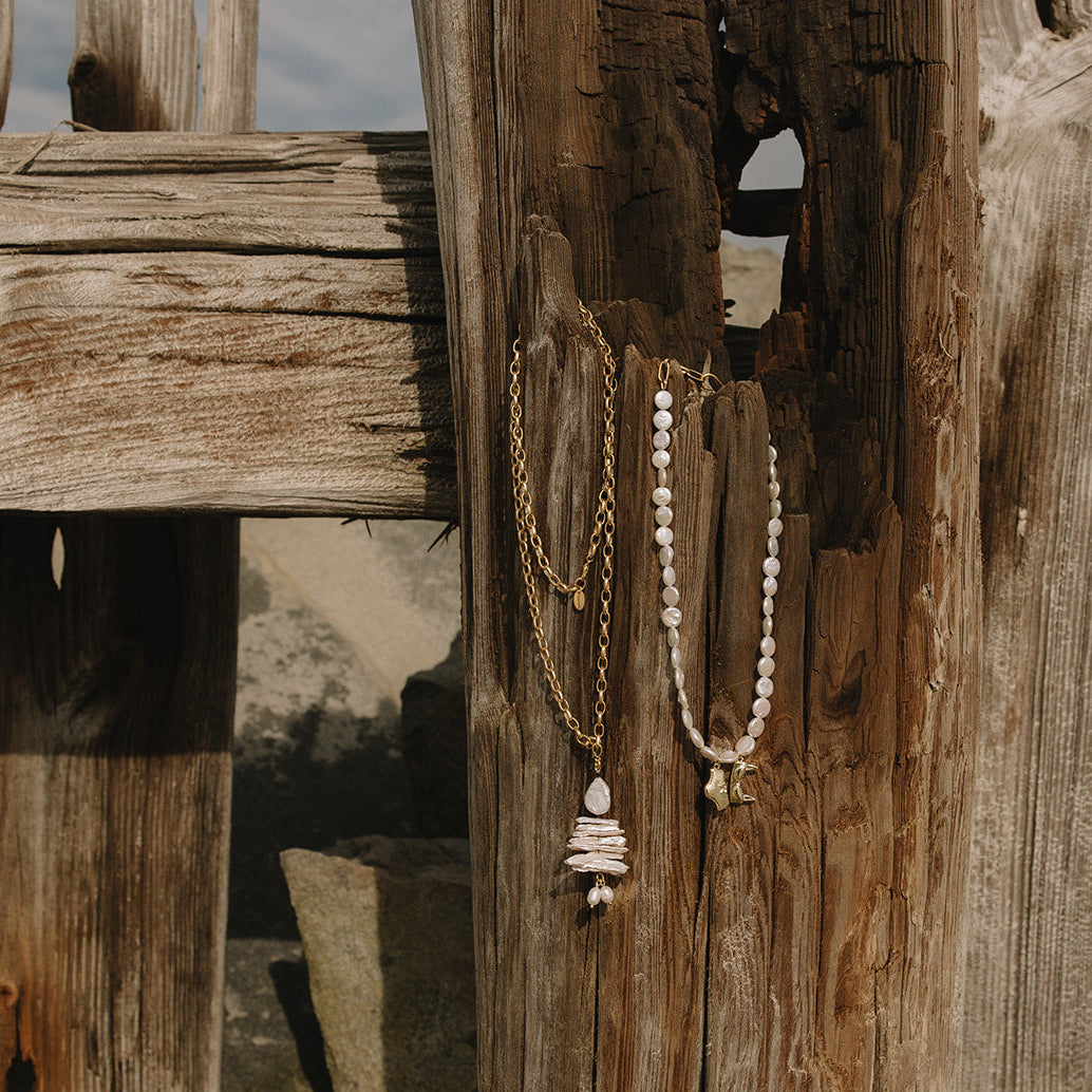 Two bohemian-style necklaces hanging from a rustic wooden fence post outdoors. One necklace has a gold chain with white pearls and a stacked white pearl pendant.