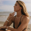 A woman sitting on a sandy beach wearing a white pearl necklace, matching pearl bracelets, and pearl drop earrings, paired with a dark bikini top.