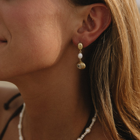 A close-up lifestyle shot of a woman wearing a delicate gold and white beaded drop earring, with a matching pearl necklace visible at the bottom.