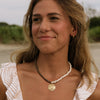 A woman with light brown hair wearing a gold pendant necklace and a white top with a beige leaf pattern, photographed outdoors against a blurred natural background.