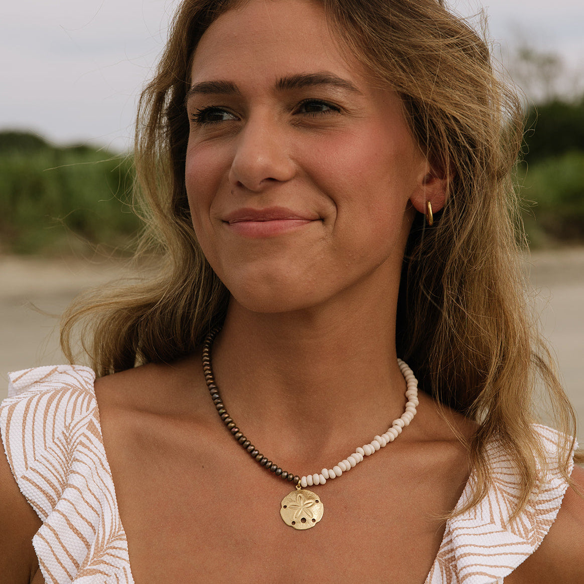 A woman with light brown hair wearing a gold pendant necklace and a white top with a beige leaf pattern, photographed outdoors against a blurred natural background.