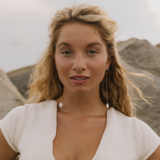A woman with blonde hair wearing a white short-sleeve knit top and long dangling earrings, photographed outdoors against a soft-focus rocky background.