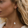 A close-up side profile of a woman wearing a delicate gold dangle earring with a white baroque pearl pendant.