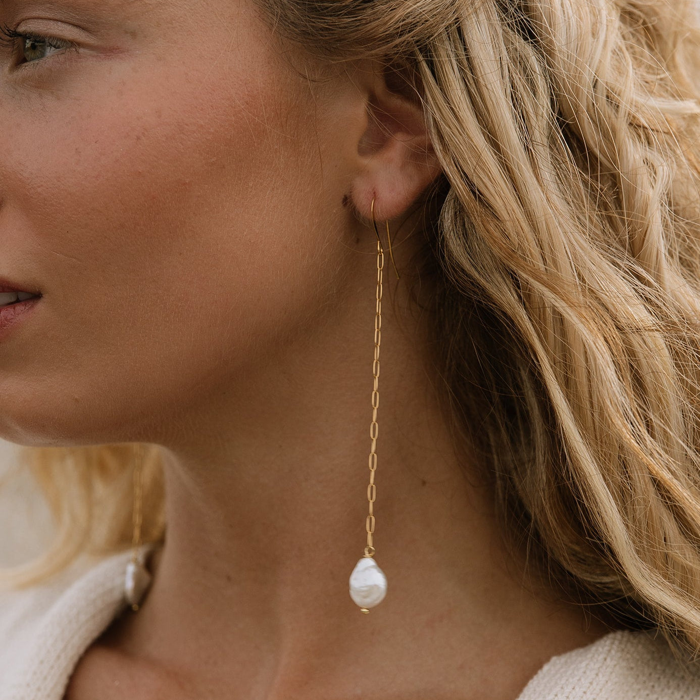 A close-up side profile of a woman wearing a delicate gold dangle earring with a white baroque pearl pendant.