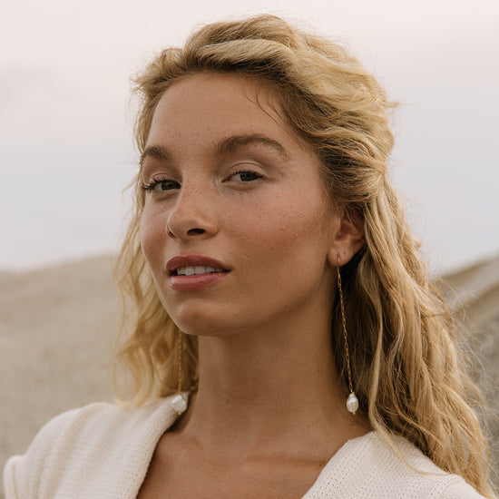 A close-up portrait of a woman with blonde hair wearing a white textured knit top and long dangling earrings, set against a soft-focus outdoor background.