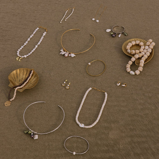 An assortment of bohemian-style jewelry pieces, including pearl and bead necklaces, gold and silver wire cuffs, and earrings, arranged on a textured brown surface with decorative seashells.