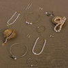 An assortment of bohemian-style jewelry pieces, including pearl and bead necklaces, gold and silver wire cuffs, and earrings, arranged on a textured brown surface with decorative seashells.