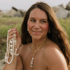 A woman outdoors wearing layered gold necklaces and bracelets with white shell-like beads, holding a strand of the beads in her hand. She is wearing a brown strapless top, with a blurred natural background of rocks and grass.