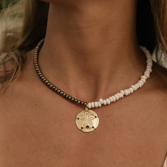 A close-up of a person's neck and chest wearing a beaded necklace with a gold-tone sand dollar pendant. One side of the necklace has small dark metallic beads, and the other side has small off-white beads, with the pendant hanging in the center.