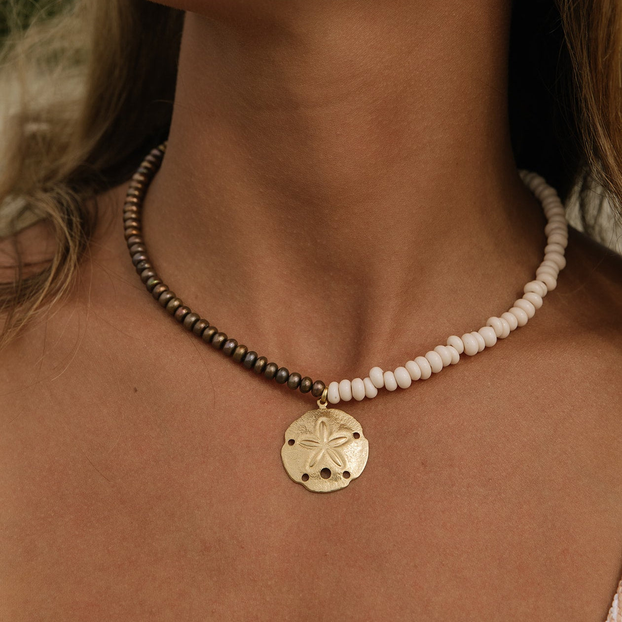 A close-up of a person's neck and chest wearing a beaded necklace with a gold-tone sand dollar pendant. One side of the necklace has small dark metallic beads, and the other side has small off-white beads, with the pendant hanging in the center.