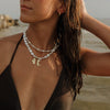 A woman on a beach wearing a black triangle bikini top and layered white shell necklaces with small gold charms.