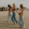 Three women walking hand-in-hand on a sandy beach, showcasing casual summer outfits including light-wash jeans, a black bikini top, and a white lace crop top.