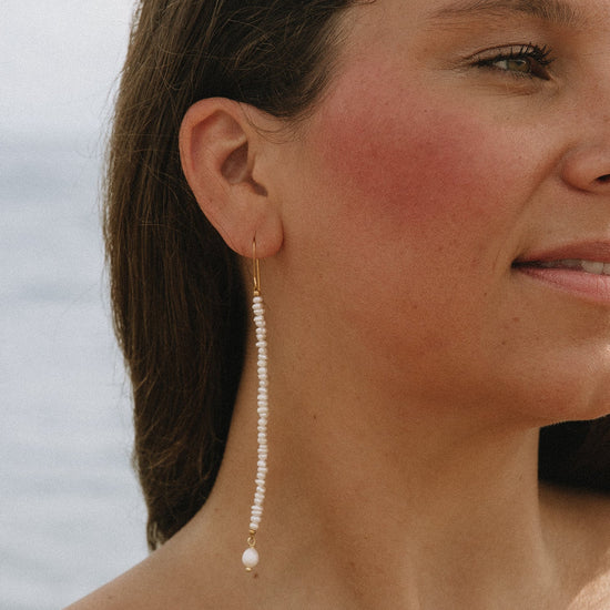 A close-up lifestyle shot of a woman wearing a long, dangling pearl earring with a gold hook and bead detailing, set against a soft, blurred background.