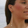 A close-up lifestyle shot of a woman wearing a long, dangling pearl earring with a gold hook and bead detailing, set against a soft, blurred background.