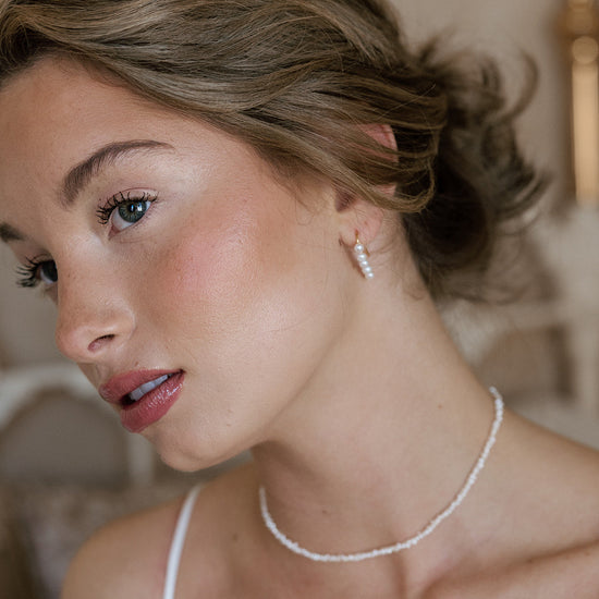 Close-up of a woman with styled hair, wearing pearl earrings and a delicate necklace. Her expression is serene, with soft lighting enhancing natural tones.